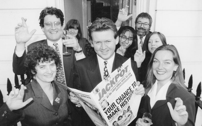 Black and white shot of editors and designers with Nick Andrews holding the Camelot newspaper 'Jackpot'. Alison Harmer, left, before she became a freelance copywriter.