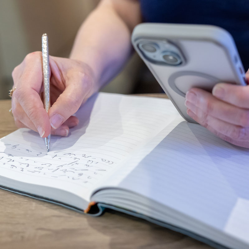 Hand of a freelance copywriter writing shorthand in a book, holding a mobile phone in the other hand