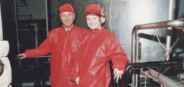 Alison Harmer with Roy Stemman in red protective boiler suits visiting a yogurt manufacturing plant, standing on a gantry, surrounded by steel pipes.